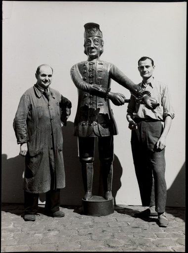 Two men on either side of metal sculpture (male figure in historical costume)