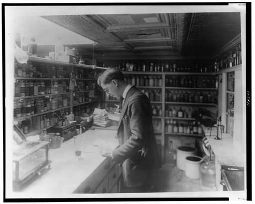 Pharmacist at People's Drug Store, No. 5, 8th and H Streets, N.E., Washington, D.C., looking at prescriptions(?) on the counter in room lined with shelves of pharmacy bottles