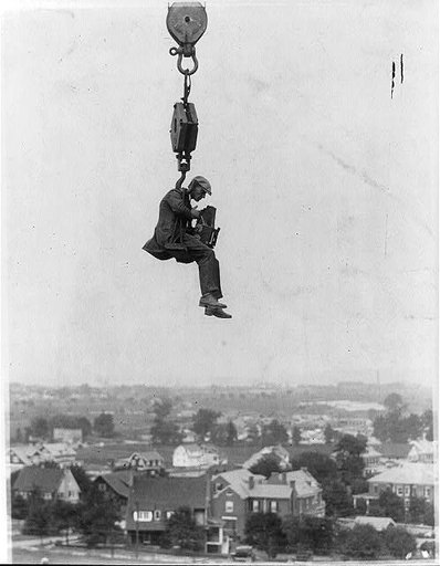 Photographer with camera suspended from hook of a (crane?) high in the air