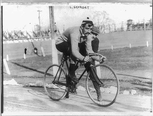 Leon Georget, full length, seated on bicycle, facing right; another man holding the bicycle