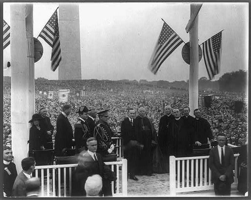 President Coolidge on speaker's stand with Army and Navy officers, and 5 Catholic priests; Washington Monument in background and large crowd on Monument grounds