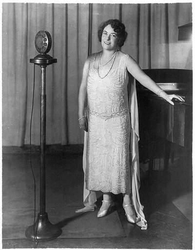 Frances Alda, full length, standing, between piano and radio microphones, facing left