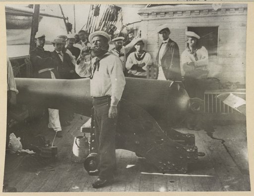 Boatswain piping in front of large cannon on deck of U.S. Naval warship; other sailors standing in background