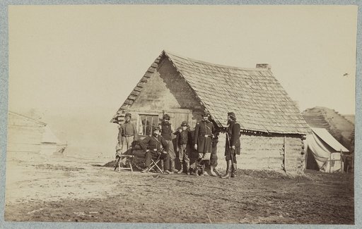 A group of soldiers, and two young men, one an African American, stand outside of log cabin quarters