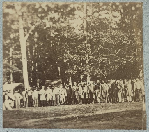 Group of soldiers, some in uniform and some with crutches, pose in front of the woods at the hospital at Camp Letterman in Gettysburg