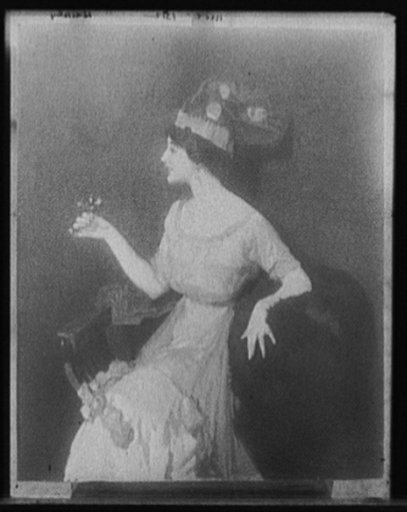 Woman holding flower, seated in chair, three-quarter length portrait