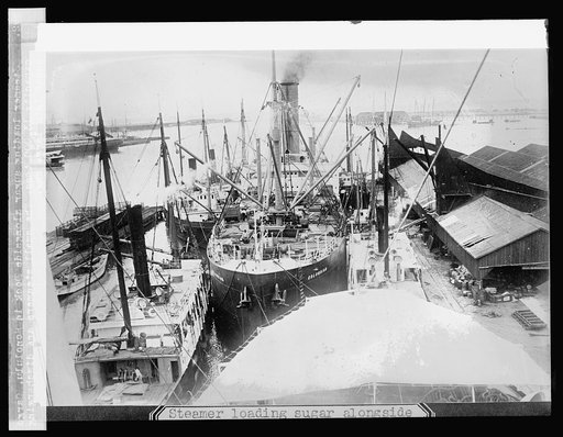 Steamer loading sugar alongside dock in Honolulu, [Hawaiian Islands]; cargo capacity 12,000 tons. The small steamers are discharging sugar into large one.
