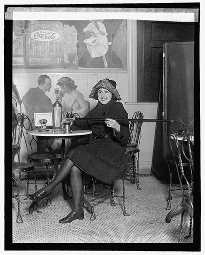 Woman seated at a soda fountain table is pouring alcohol into a cup from a cane, during Prohibition; with a large Coca-Cola advertisement on the wall], 2/13/22