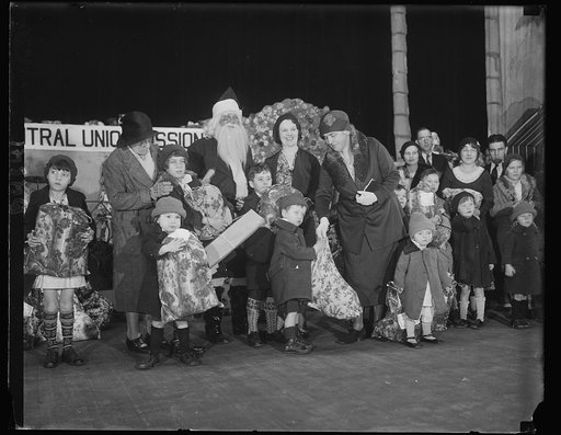 First Lady assists Santa. Mrs. Hoover took an active part in the Christmas affair for Washington's less fortunate residents. She is shown handing out one of the 1,500 bags of food and presents which were distributed by the Central Union Mission at a party held in the Fox Theatre. Assisting Mrs. Hoov