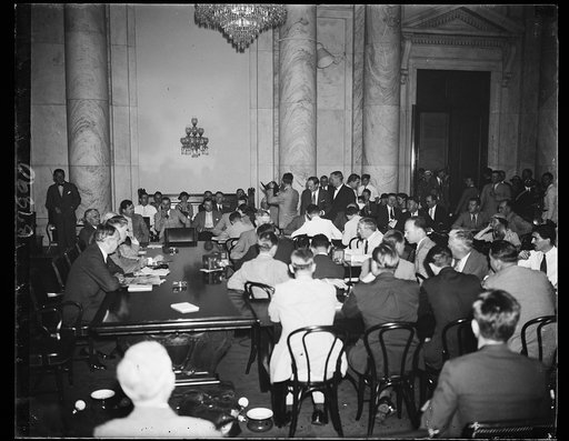 Bombardment starts. On the right of the table, profile showing, is Ewing Y. Mitchell, ousted Assistant Secretary of Commerce, appearing before the Senate Commerce committee to tell his charges of graft in the Commerce Department. On the left are Senators Arthur Vandenberg, R. of Mich., and Royal S. 