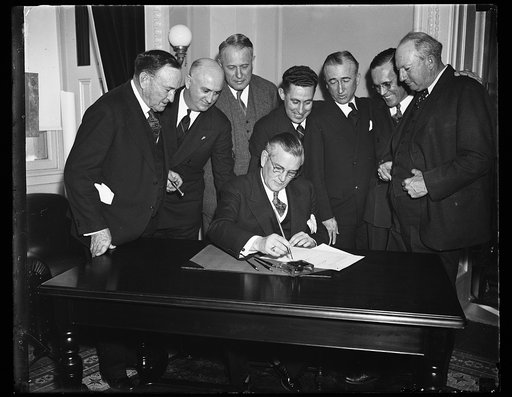 Man signing document at desk; group includes Joseph T. Robinson, left; Pat Harrison, right