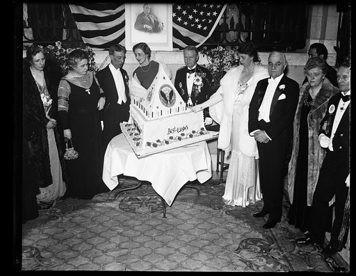 FIRST LADY LIGHTS BIRTHDAY CAKE. MRS. ROOSEVELT LIGHTS THE CANDLES ON THE BIRTHDAY CAKE AT THE SHOREHAM HOTEL BIRTHDAY BALL OF PRESIDENT ROOSEVELT. THE PRESIDENT REMAINED AT THE WHITE HOUSE AND HAD WITH HIM A FEW CLOSE FRIENDS. IN THE ABOVE PICTURE MRS. WOODROW WILSON IS ON THE EXTREME LEFT. DIRECTL
