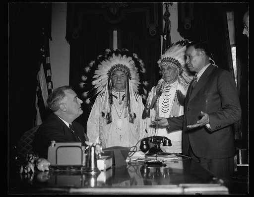 INDIANS VISIT GREAT WHITE FATHER. PRESIDENT ROOSEVELT, SEATED, RECEIVES DRAGS WOLF, LEFT, AND FOOLISH BEAR, RIGHT; WHILE ARTHUR MANDAN, CHIEF OF THE TRIBAL COUNCIL, RIGHT; INTERPRETS FOR THE INDIANS. THE PURPOSE OF THE VISIT WAS TO THANK THE GREAT WHITE FATHER FOR HIS HELP IN RESTORING THE 'SACRED B