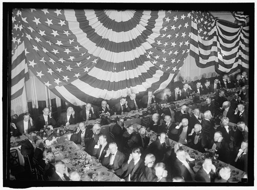 Banquet scene with flag draping and men seated holding up listening device to one ear