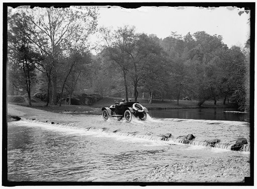Couple in automobile fording a river at a low water crossing near a small stone bridge