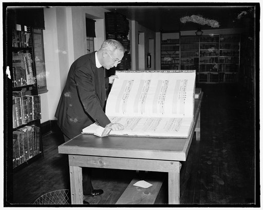 Man in clerical collar looking at oversize book with musical score, possibly Handel's "Drxit Dominus," in a library
