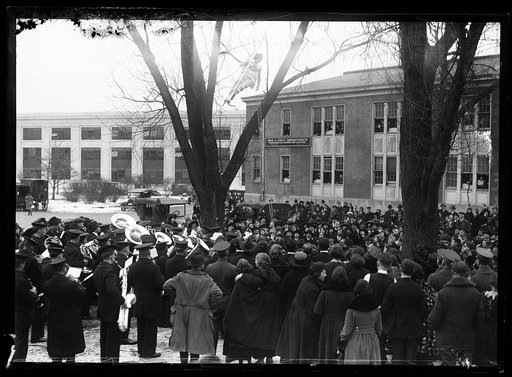 Crowd watching acrobat suspended in air near building for War and Navy Departments, Commissions on Training Camp Activities. Washington, D.C.