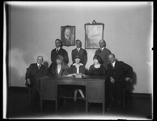 Group seated at desk with portraits of Herbert and Lou Hoover on wall
