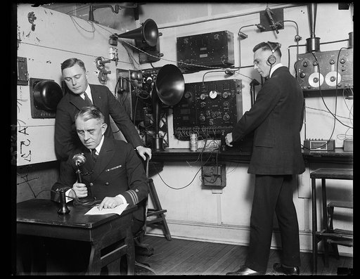 Hdqtrs. of the U.S. Coast Guard are using unique method for distributing news of their service by means of radio. Capt. F.C. Billard, aide to the commandant is shown broadcasting. Lt. F.A Zensler is coaching him from the receiving set which is being used by Lt. E.M. Webster (with ear pieces)