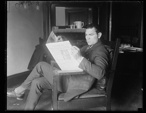 Boxer Jack Dempsey seated in a chair, reading a newspaper