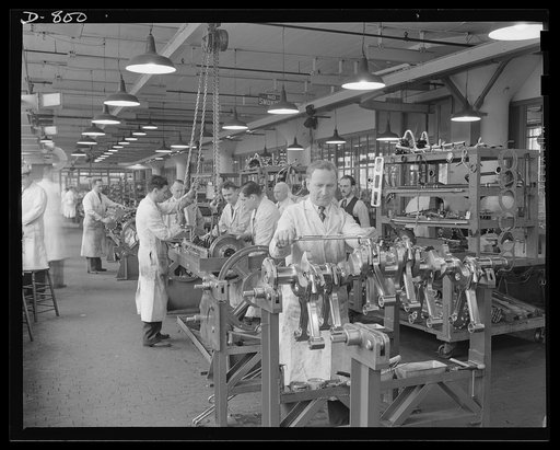 General view of engine assembly line, Packard plant, Detroit, Michigan. Man in foreground is checking tension on connecting rod bolt nuts. The men at the bench behind him are assembling the crankshaft and connecting rod assembly, and installing them in the engine. Packard marine engine plant