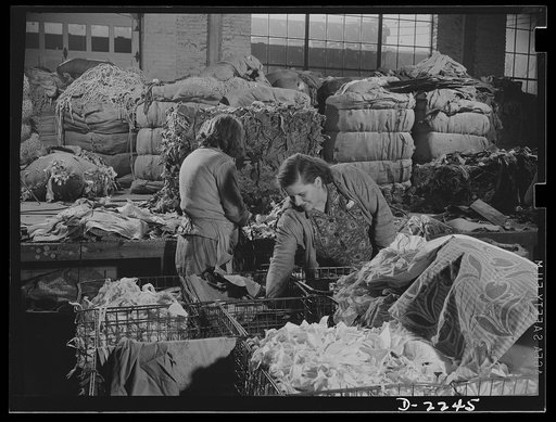 Rags. Collection and processing. A "team" of two women sorting rags. In the background are a number of bales waiting to be sorted. On the table behind the woman at left is the remainder of a bale which she has been sorting. In the foreground are baskets containing the rags that have been sorted. Sha