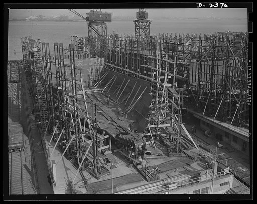Shipbuilding. "Liberty" ships. A new vessel has just moved off these ways, and the keel is being assembled for yet another in a large Eastern shipyard. The bottom and side shell plating and the inner tank sections are being assembled. All parts are prefabricated in this huge Eastern plant which form
