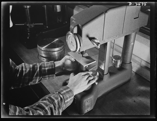Conversion. Copper and brass processing. This sample of brass strip is being tested on a Rockwell Hardness Testing Machine. This test is based upon the depth a steel ball penetrates the metal. A minor load is applied first to hold the ball in position, and then a major load is applied. The dial on t