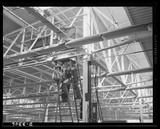 A big auto plant changes over to the production of machine guns for the Air Force. Electricians begin running power lines to the new machine locations. Oldsmobile, Lansing, Michigan