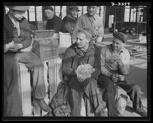 Conversion. Heaters to bogie wheels. How'd it go today with the boys at Bataan? These employees of a Midwest heater company take time out for lunch and the news before resuming work on installing assembly apparatus for M-4 tank bogie wheels in a company warehouse