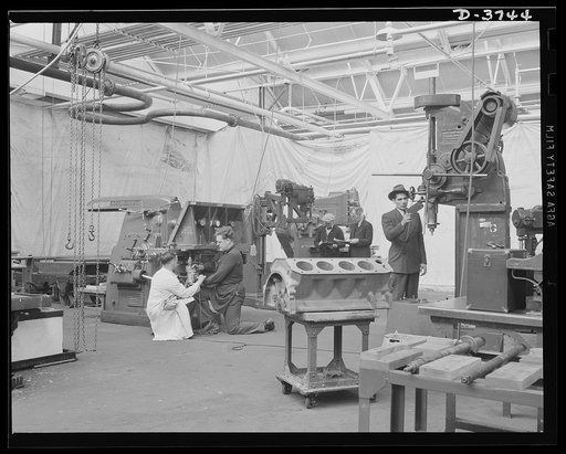Preparing for tank production. A temporary machine shop is set up in a big automobile plant. A large drill press , a radial drill press, a vertical mill and a horizontal mill are background for an engine block of the type to be used in the tank motor. Ford Lincoln plant, Michigan