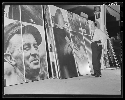 The artist inspects sections of the huge Arsenal of Democracy photomontage, which, when assembled, will be 15 x 30 feet. The artist is Jean Carlu who designed this panel and one portraying the Four Freedoms for exhibition by the Office of Emergency Management (OEM) at the National and Civilian Defen