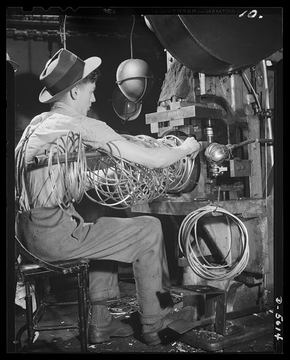 Conversion. Auto radiators to army helmets. The rim of an army helmet is formed on a press from steel rod. Finished rims are shown on the hook below the operator's right hand. The helmets are made in an Eastern plant converted by retooling from the production of automobile radiators. McCool Radiator
