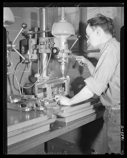 Conversion. Paper machinery to plane wing parts. A drill press operator bores holes in an airplane wing hinge in the plant of an Eastern paper machinery manufacturer. The hinge is clamped to the under side of a drill jig. The company also makes naval sights and other war essentials