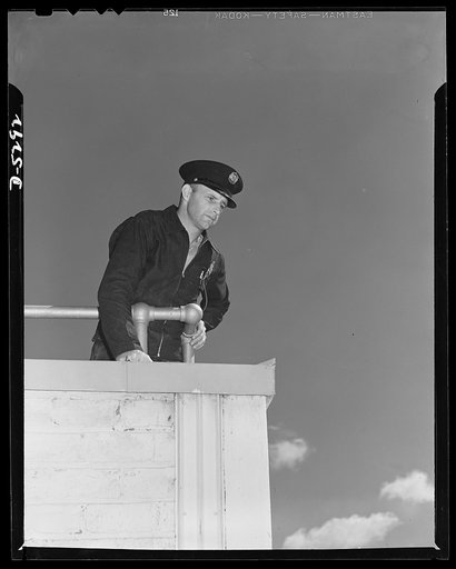 Production. Aircraft. High above the roof of a Western aircraft plant covering nearly two million square feet of shop and assembly space, this fireman keeps a watchful eye on the scene below. During an air raid his post is here, as a spotter for the incendiary's flare