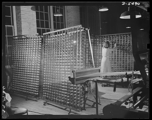Production. Parachute cloth and shrouds. A warping operation in the making of parachute cloth and shrouds. The spools of silk are put on racks and drawn through a big reel. From there they go to a roll for weaving