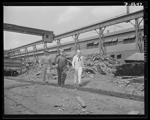 A poster comes to life. The three men tour one of the mill yards. In the background, to the immediate right of the freight car, is a portion of a scrap pile. Immediately behind Sergeant Vineyard is a pile of limestone and slag. In the background, to the sailor's right, are piles of iron heaped high.