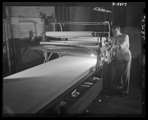 Production. Parachute making. Rolls of silk, destined for action as parachutes for the U.S. Air Force, shown here on the laying-out and cutting table of an Eastern manufacturing plant. The folder machine folds layer upon layer of silk for the cutting operation. Pioneer Parachute Company, Manchester,
