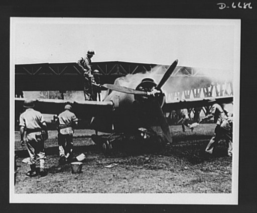 Marines in combat. Fire in this fighter plane was quickly extinguished by Marines at the Guadalcanal airport after a Japanese bomb hit the hangar shown in the background. The plane was not seriously damaged