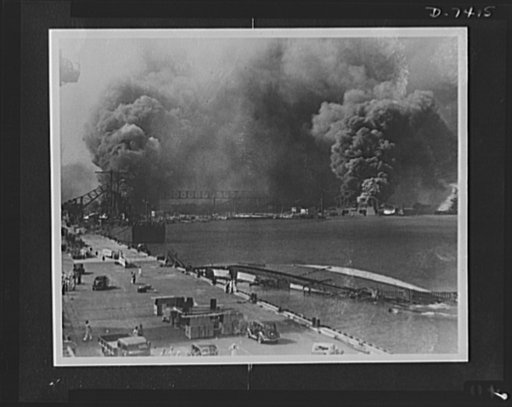 Pearl Harbor bombing. Destruction. Smoke pours from the USS Shaw, bombed dry dock (right center) while in the foreground lies the capsized USS Oglala, a minelayer. To the left is the 10,000 ton cruiser, USS Helena, struck by an aerial torpedo on the starboard side. The concussion caused the Oglala, 