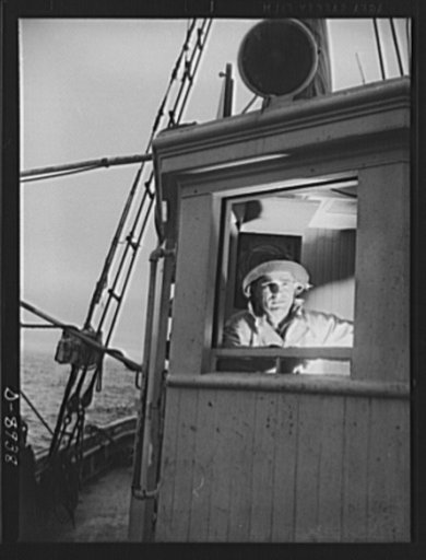 Victory food from American waters. With the light from the compass binnacle illuminating his face, Captain John Riberia [i.e., Ribeiro] guides Old Glory back to Gloucester port. Decks have been cleared and fish stored in ice until return to port