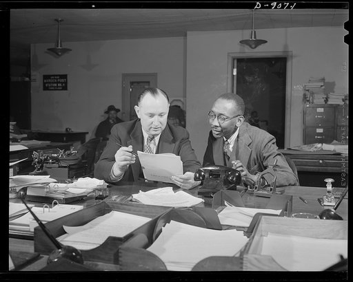 Office of War Information news bureau. Ted Poston, shown here with Elmer Roessner, chief of the general news desk, sees that Negro papers across the country get their share of war news. Tale of a City, an OWI pamphlet on Nazi treatment of Poland, which Poston and Roessner are discussing here, was di