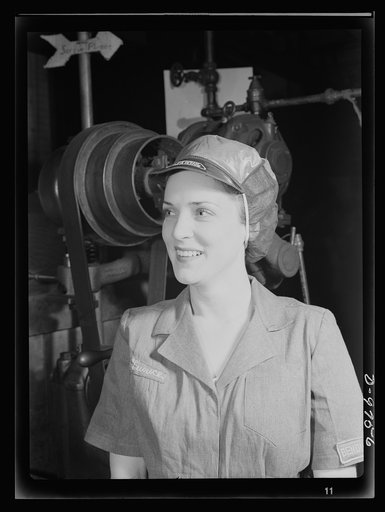 Safe clothes for women war workers. Her hair tucked away under a safety cap, war worker Eunice Kimball stands beside whirling machine wheels with no wisp of hair exposed. She's completely protected from the danger of entanglement in moving machine parts. Bendix Aviation Plant, Brooklyn, New York