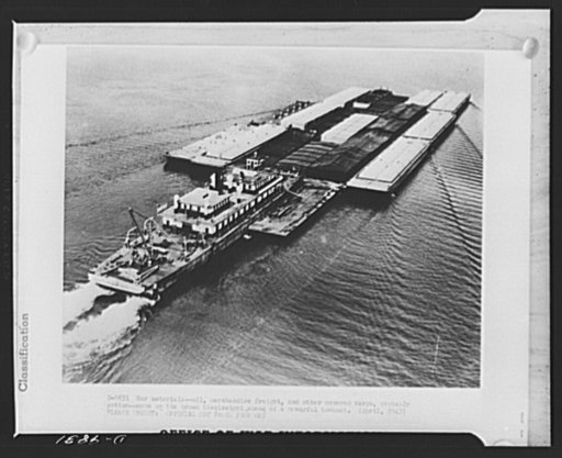War materials. Oil, merchandise freight, and other covered cargo, probably cotton, move up the broad Mississippi ahead of a powerful towboat