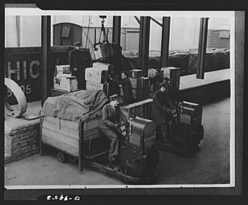 Lend-lease to Britain. Cases of American spare parts arriving at an English ordnance center as part of a lend-lease shipment from the United States are moved to the stores for unloading. English girls, driving trucks, help with the task