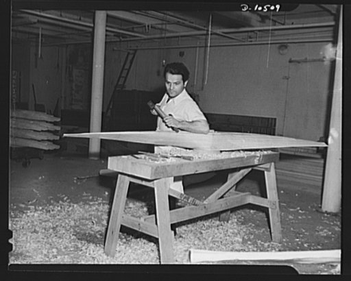 Peacetime sculptor, artist, and wood carver chiseling pitch of laminated propeller blade; two other employees of propeller department are ornamental wood carvers