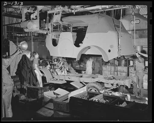Army truck manufacture (Dodge). The body of a Dodge Army reconnaissance car being lowered into place on the assembled chassis. Bodies for each of the many types of army trucks are synchronized in production with the chassis for which they are intended and are carried from a distant part of the plant
