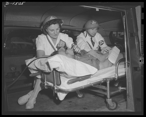 Civilian protection. No time to spare before treating this patient, so these members of the emergency medical aid corps (see armbands insignia) go to work on a bombing victim right in the ambulance. The young lady is operating an inhalator to keep the victim breathing, while the doctor (note the M.D