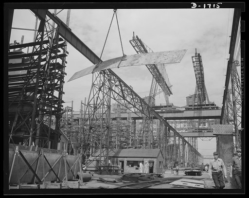 Shipbuilding (Newport News). A huge overhead crane swings this metal plate into position to be welded to a new cruiser under construction for the U.S. Navy