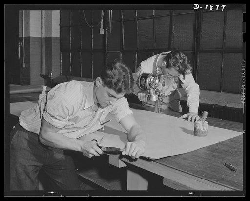 Barrage balloon manufacture. Neck and neck in the stretch. Appearing like race horses galloping down each side of a long strip of barrage balloon fabric, the worker with the scissors apparently finishing ahead of the fellow with the electric fabric cutter. Actually the scissors are not used for cutt